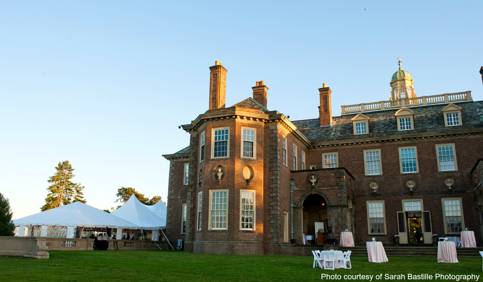 View of Estate with cocktail tables in front and peaked tent on the side of building. 