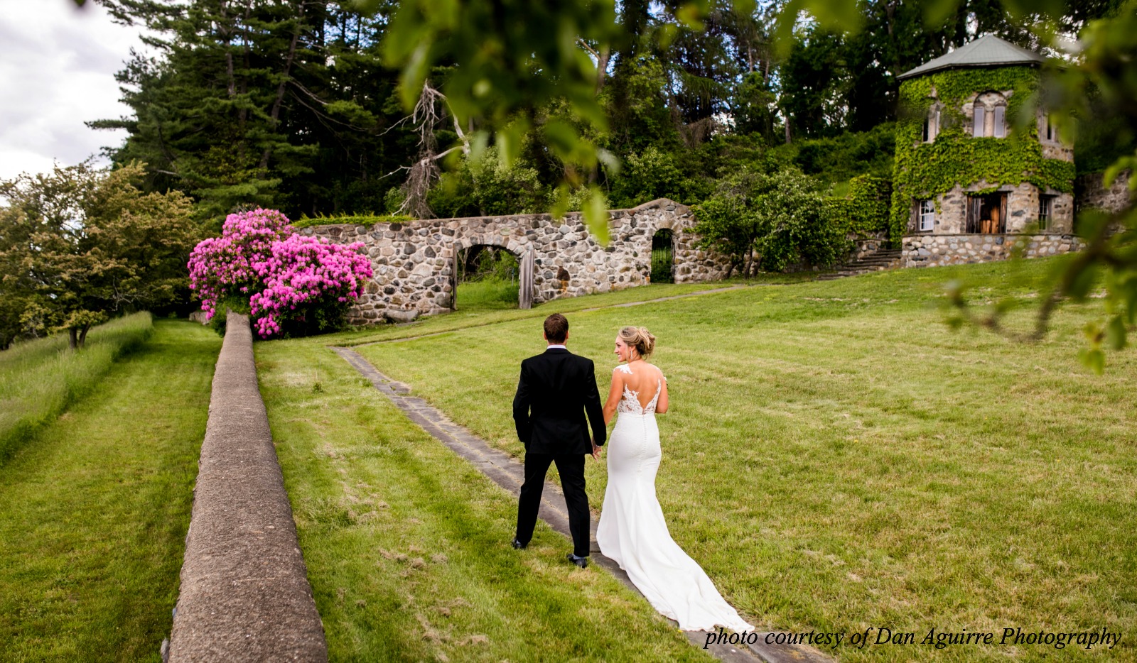 Bride and Groom walking along garden near The Barn at The Crane Estate along stone walls. 