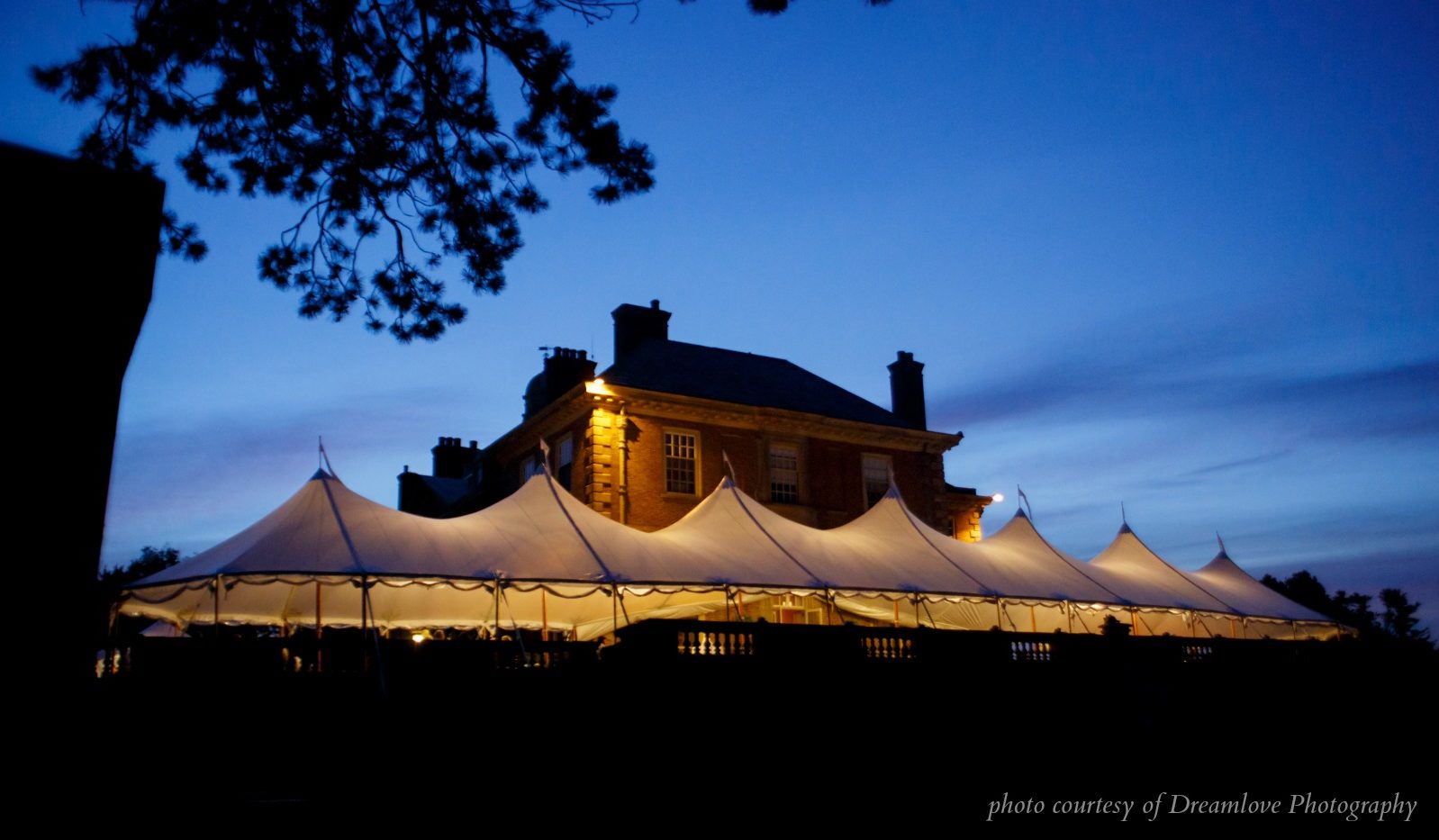 Large tent with six peaks, evening view next to estate, tent aglow. 